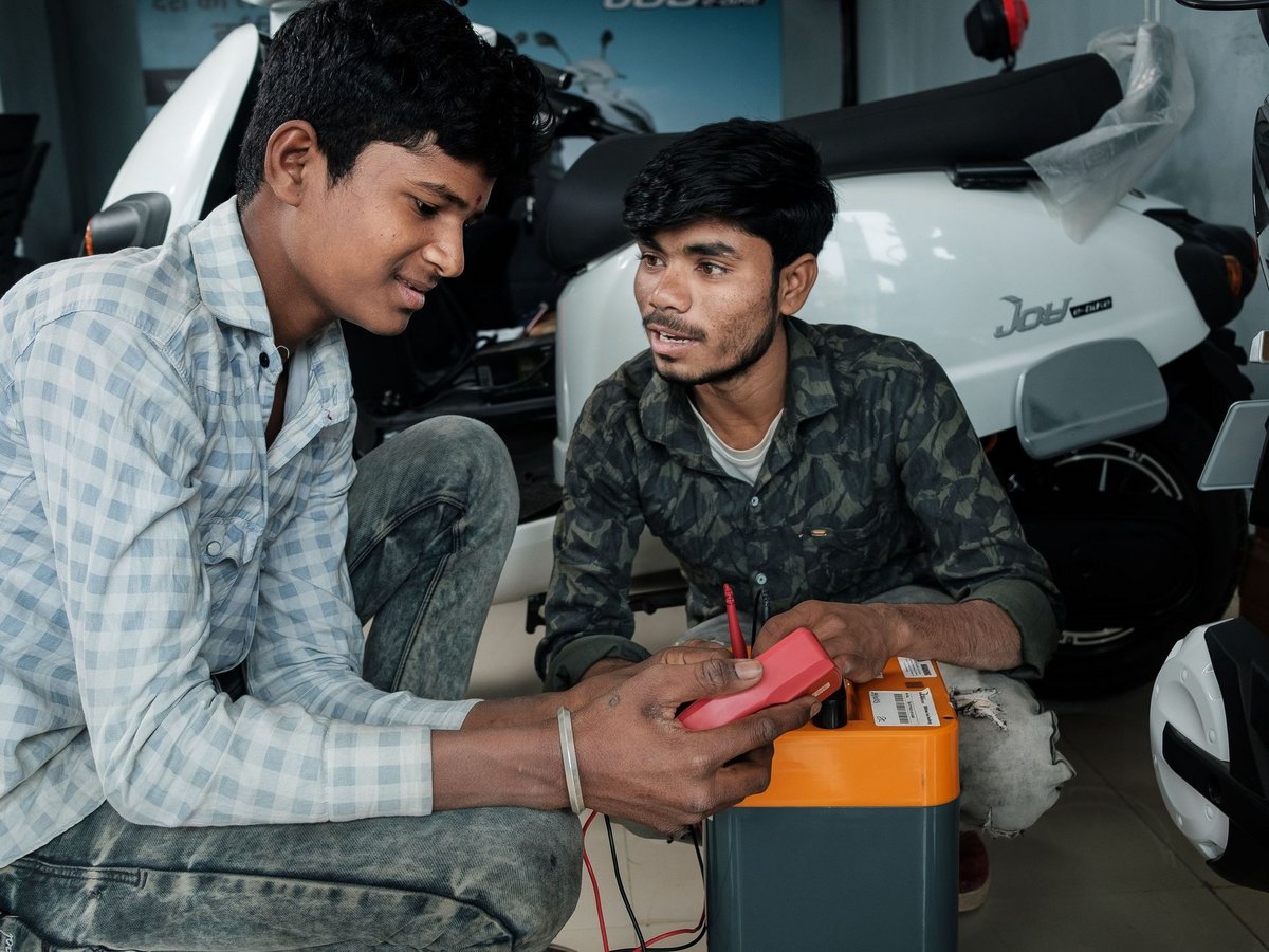 Zwei junge Männer prüfen eine Elektrorollerbatterie mit einem Multimeter in einer Werkstatt, umgeben von E-Bikes.