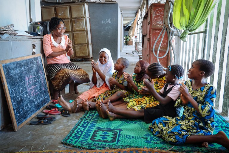 Eine erwachsene Frau unterrichtet lächelnde Kinder auf einer Matte mit einer Tafel, die eine Französischlektion zeigt.