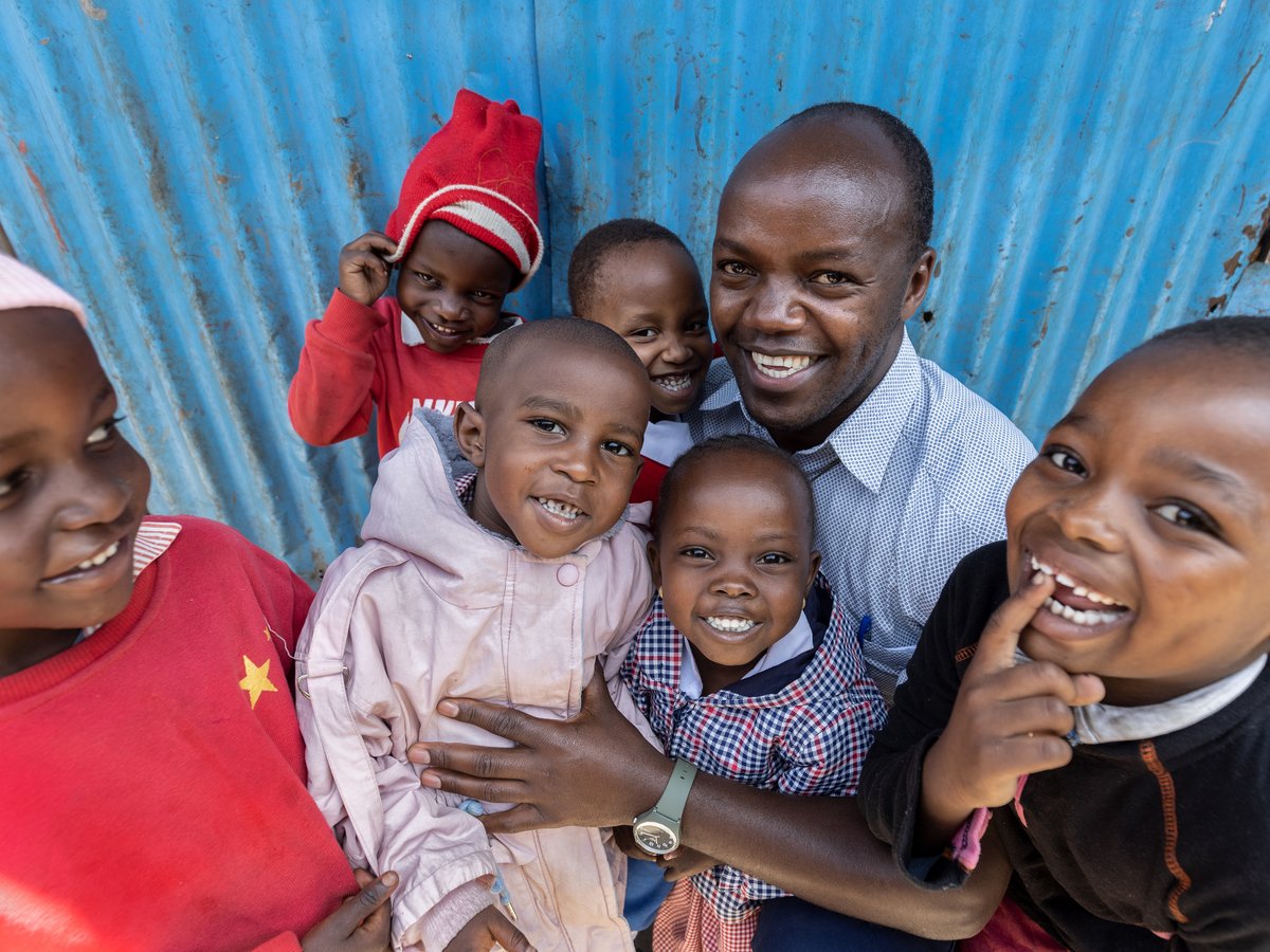 Ein lächelnder Mann umarmt mehrere lachende Kinder vor einer blauen Wellblechwand.