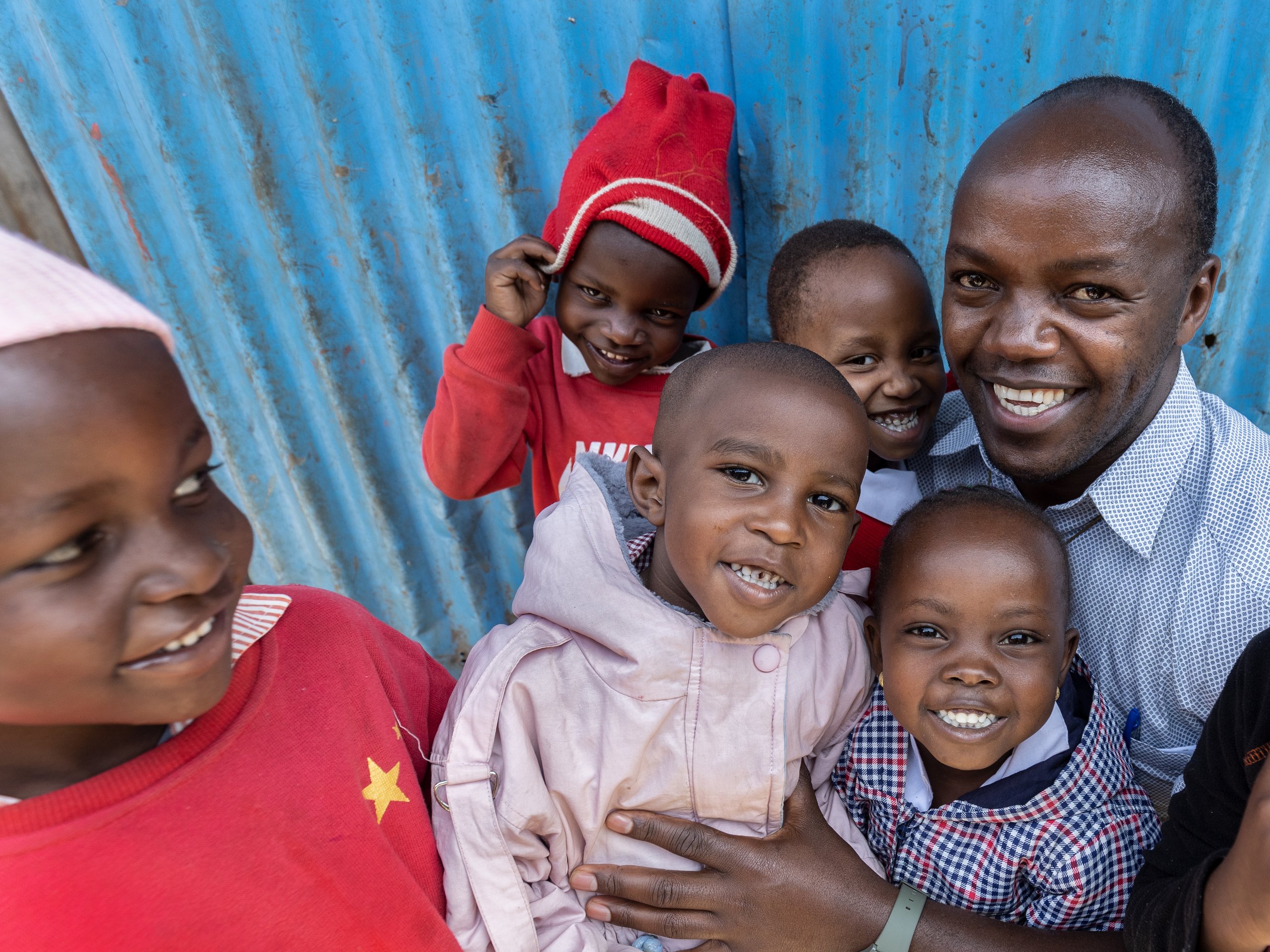 Ein lächelnder Mann umarmt mehrere lachende Kinder vor einer blauen Wellblechwand.