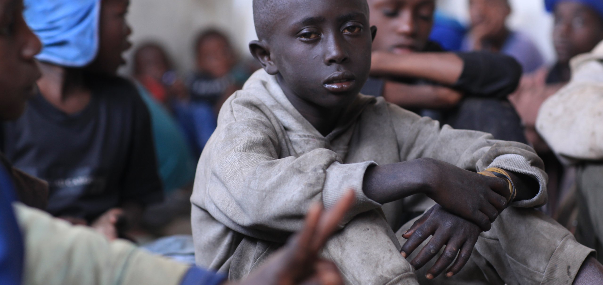 Ein ernster kleiner Junge in einem hellgrauen Kapuzenpullover sitzt zwischen anderen Kindern drinnen und blickt den Betrachter direkt an.