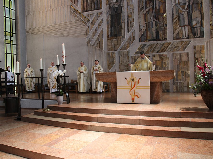 Ein Priester erhebt die Monstranz am Altar während eines Gottesdienstes, mit Klerus, Kerzen und moderner Kunst im Hintergrund.