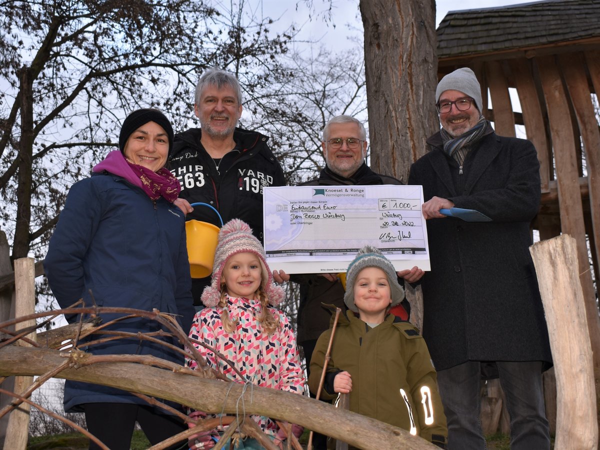 Gruppe lächelnder Erwachsener und zweier Kinder mit 1.000-Euro-Spendenscheck für Don Bosco Würzburg im Freien im Winter.