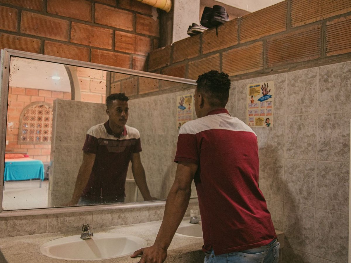 Junger Mann in rot-weißem Hemd blickt in sein Spiegelbild in einem alten Spiegel über Waschbecken in rustikalem Raum.