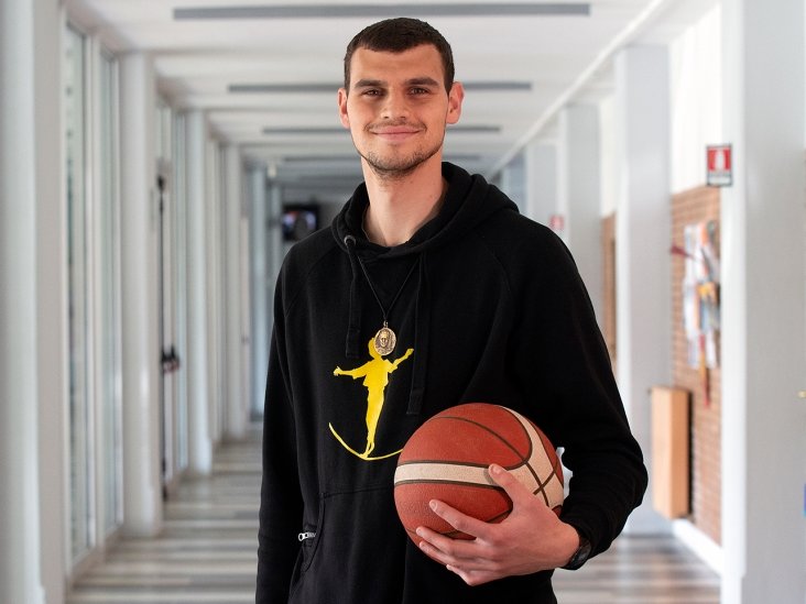 Lächelnder junger Mann in einem schwarzen Kapuzenpullover, der einen Basketball in einem modernen Flur hält.