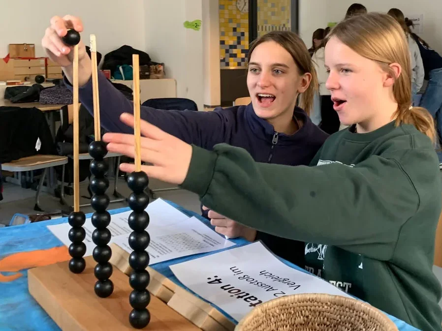 Zwei Mädchen beschäftigen sich enthusiastisch mit einem Zählgerät aus Perlen in einem Klassenzimmer, wohl bei einer Klima-Aktivität.