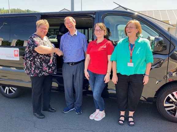 Fahrzeugübergabe: Vier Personen lächeln neben einem schwarzen Caritas-Bus des Caritasverbandes Westeifel e.V., zwei schütteln Hände.