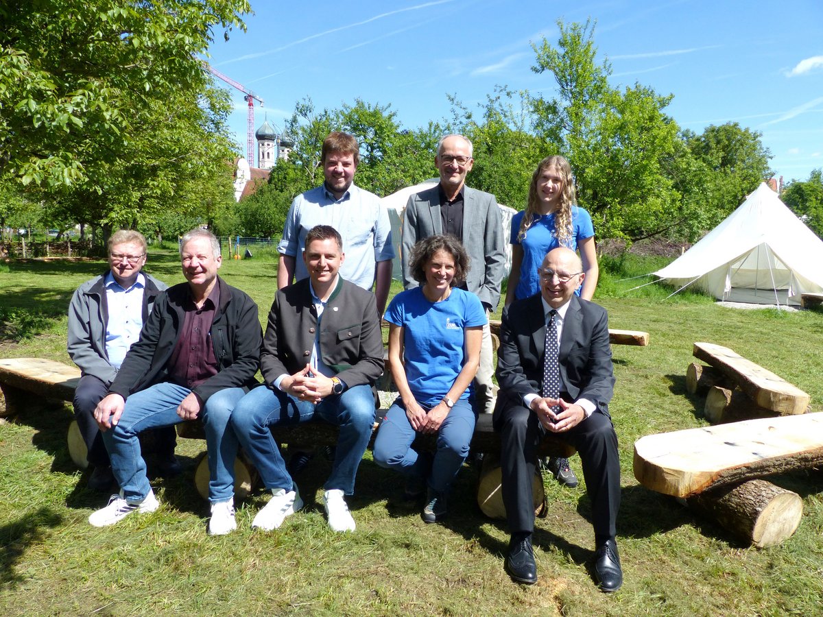 Acht Personen, teils in Tracht oder Anzügen, sitzen auf Holzbänken im Freien, mit Kirche und Zelt im Hintergrund.