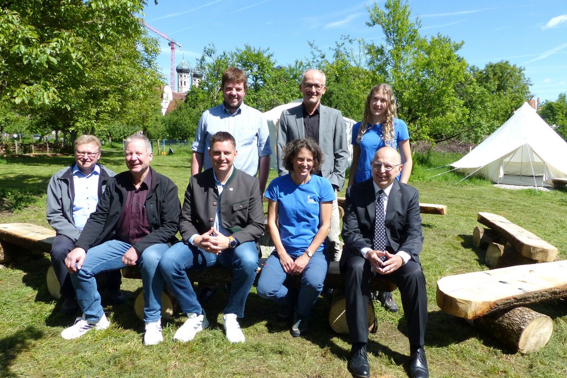 Acht Personen, teils in Tracht oder Anzügen, sitzen auf Holzbänken im Freien, mit Kirche und Zelt im Hintergrund.
