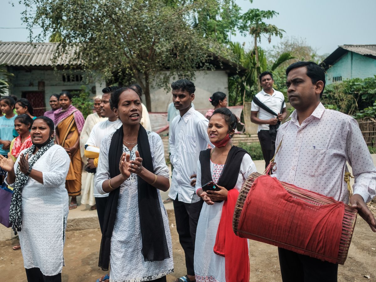 Eine Gruppe von Menschen im Freien, Frauen klatschen, eine spricht, und ein Mann hält eine traditionelle rote Trommel.