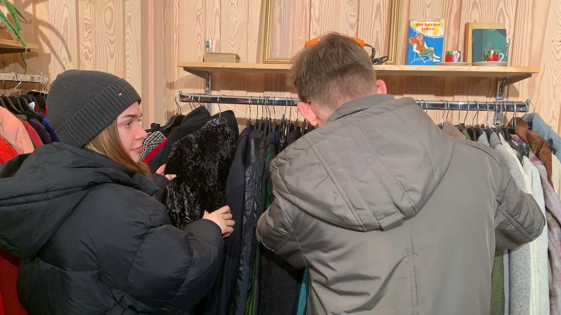 Eine Frau und ein Mann stöbern in Kleidung an einem Kleiderständer in einem Vintage-Laden mit Holzwand.