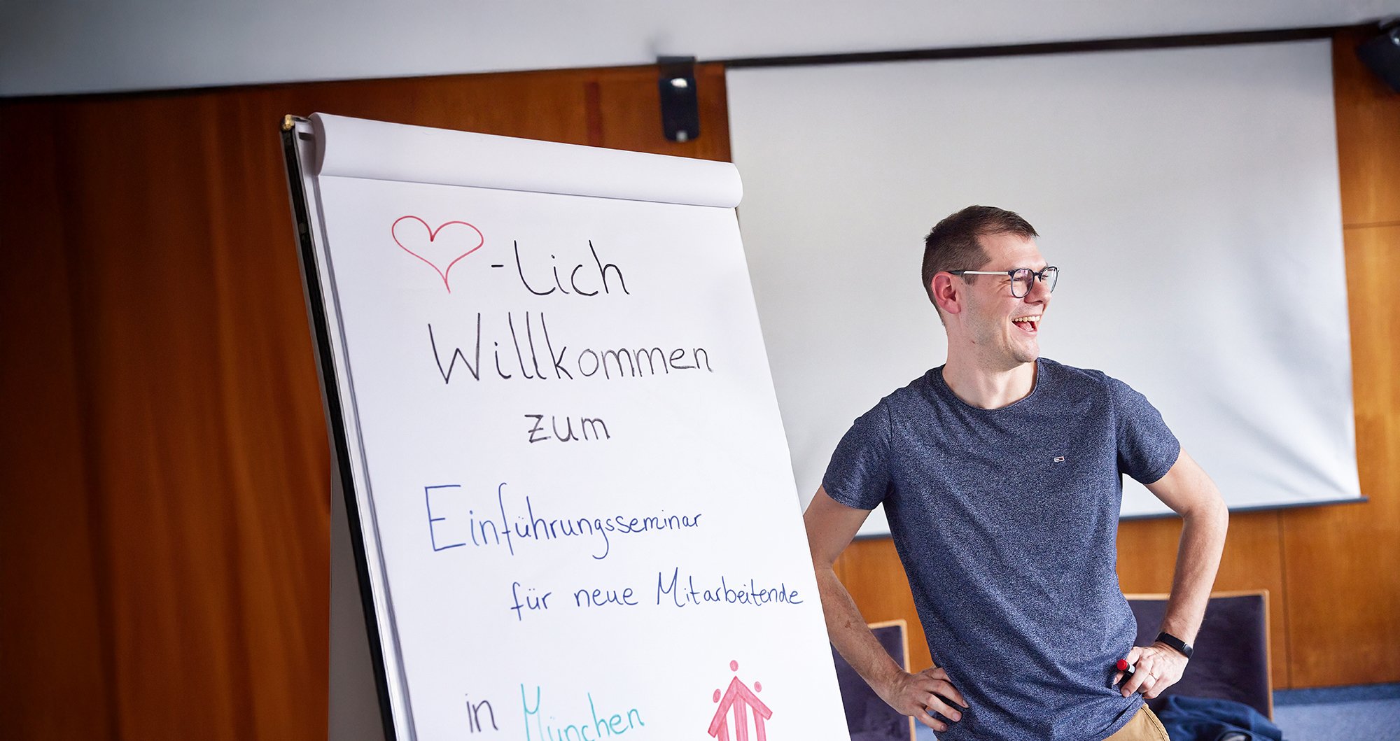 Lachender Mann steht neben Flipchart mit der Aufschrift 'Willkommen zum Einführungsseminar für neue Mitarbeitende in München'.