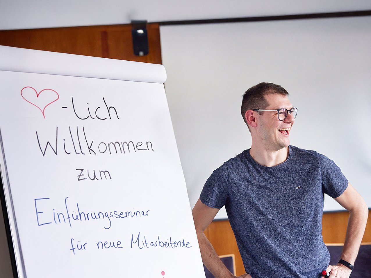 Lachender Mann steht neben Flipchart mit der Aufschrift 'Willkommen zum Einführungsseminar für neue Mitarbeitende in München'.