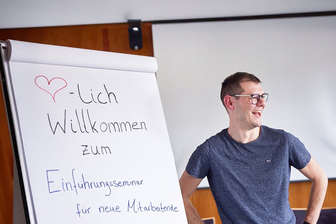Lachender Mann steht neben Flipchart mit der Aufschrift 'Willkommen zum Einführungsseminar für neue Mitarbeitende in München'.