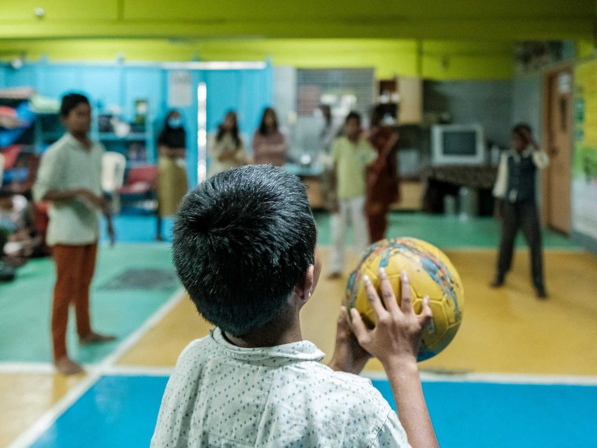 Ein Kind von hinten hält einen gelb-blauen Ball und blickt in einen Raum mit anderen Menschen.