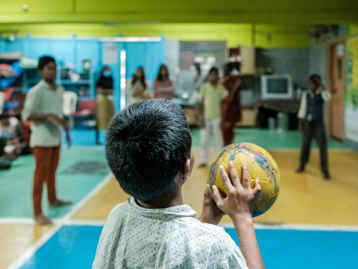 Ein Kind von hinten hält einen gelb-blauen Ball und blickt in einen Raum mit anderen Menschen.