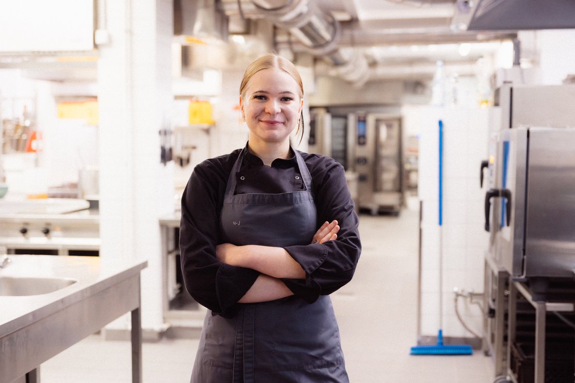Lächelnde Köchin mit blonden Haaren und Schürze steht mit verschränkten Armen in einer professionellen Küche.
