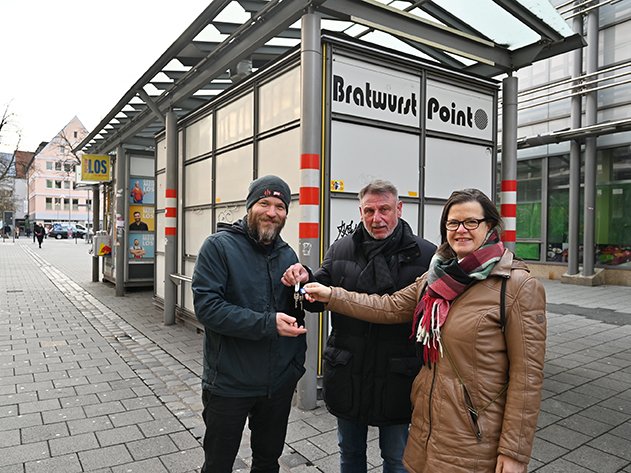 Drei Personen auf einer Stadtstraße, eine übergibt einem anderen Schlüssel, vor einem „Bratwurst Point“-Stand.