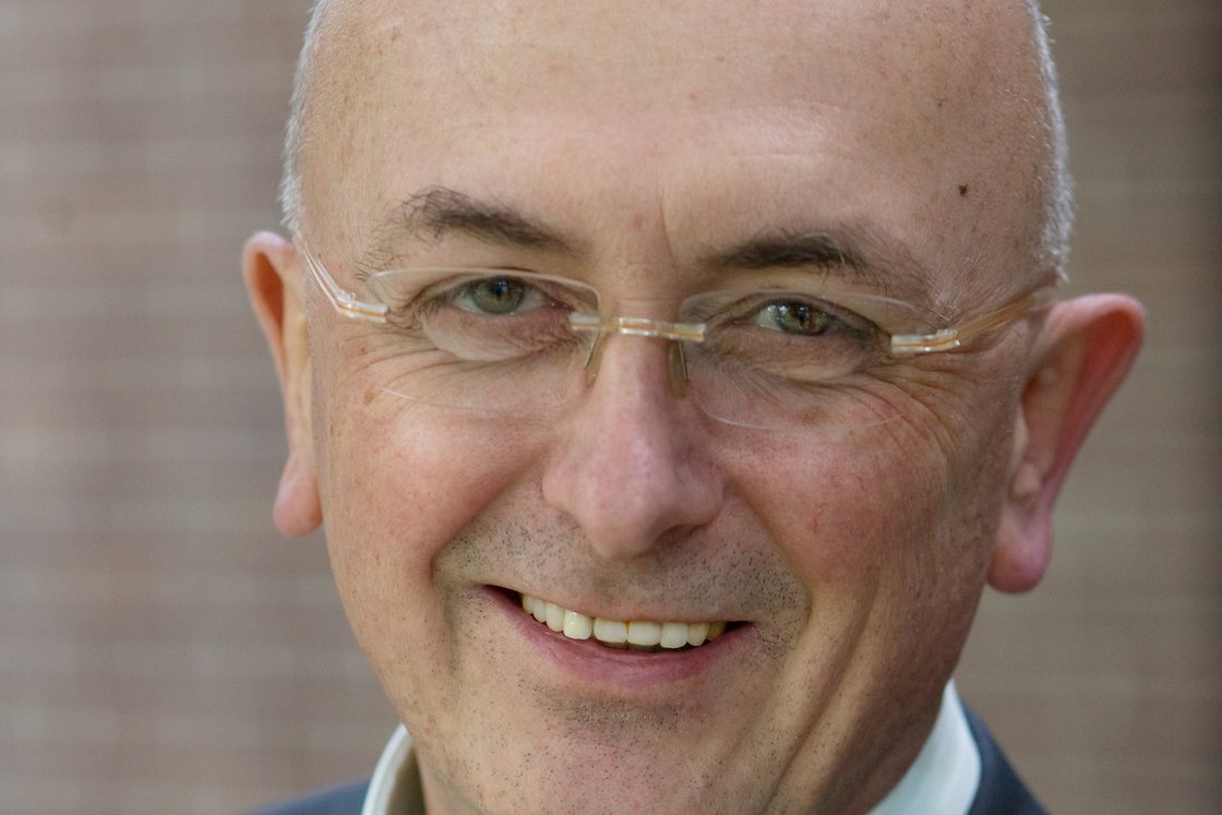 Ein lächelnder, kahler Mann mit Brille, hellgrünem Hemd und dunklem Anzug im Nahaufnahme-Porträt vor einer Ziegelwand.