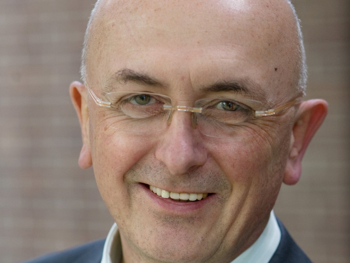 Ein lächelnder, kahler Mann mit Brille, hellgrünem Hemd und dunklem Anzug im Nahaufnahme-Porträt vor einer Ziegelwand.