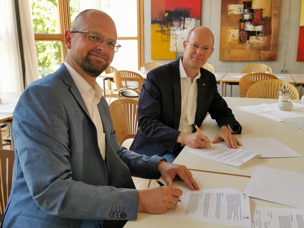 Zwei Männer in Geschäftskleidung unterschreiben Dokumente an einem langen Tisch. Einer lächelt in die Kamera, der andere schreibt.
