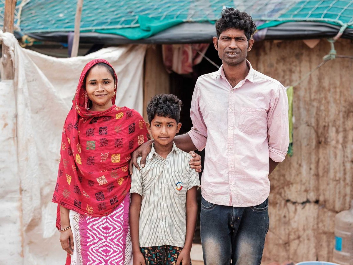 Eine lächelnde dreiköpfige Familie (Frau, Mann, Junge) steht vor einem einfachen Haus.