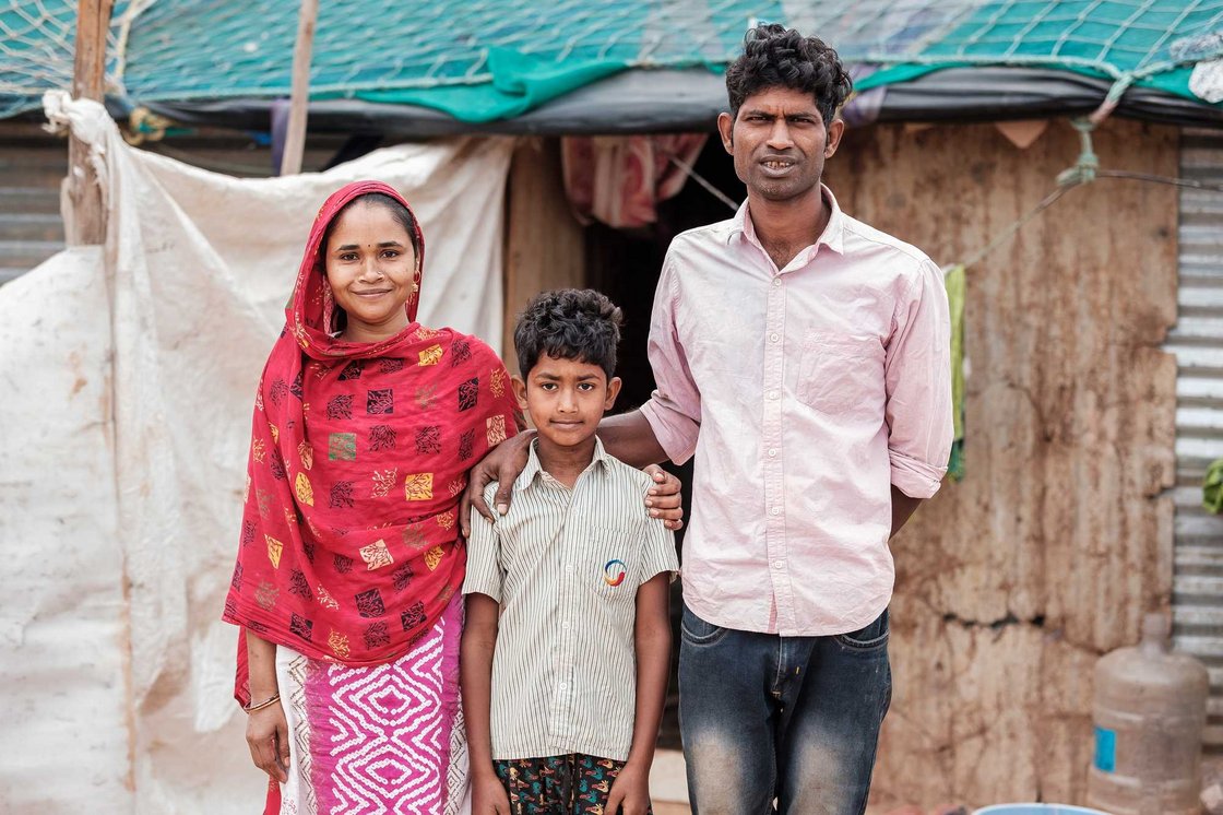 Eine lächelnde dreiköpfige Familie (Frau, Mann, Junge) steht vor einem einfachen Haus.