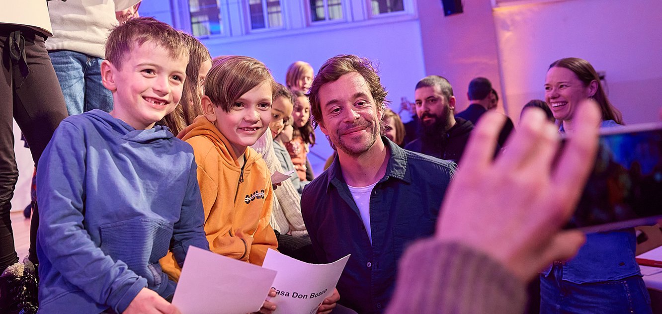 Chekcer Tobi sieht in eine Handykamera, daneben sitzen Kinder