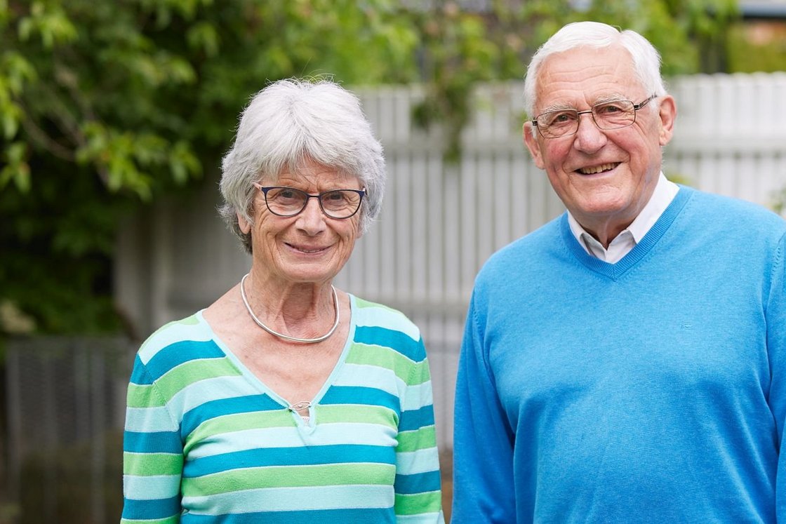 Lächelndes Seniorenpaar mit grauem Haar und Brille steht draußen.