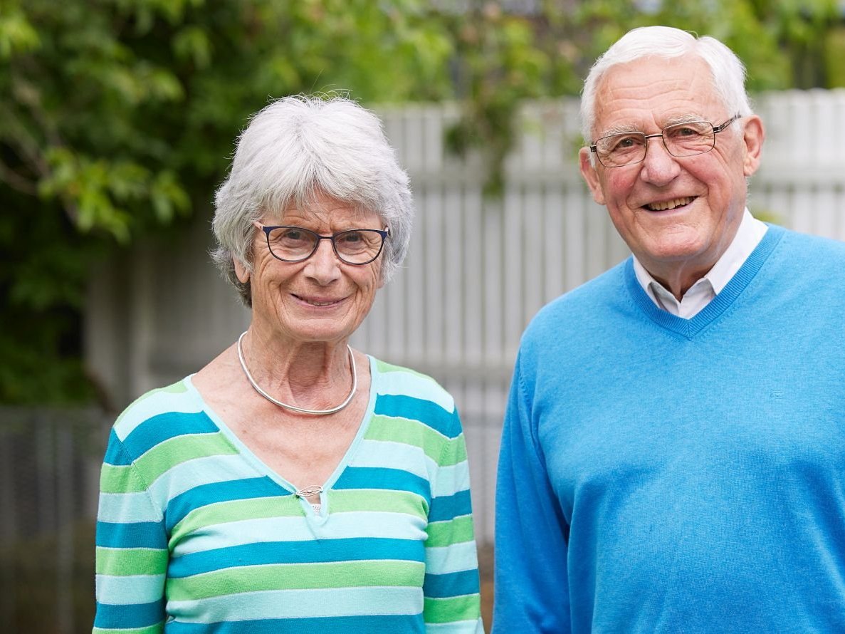 Lächelndes Seniorenpaar mit grauem Haar und Brille steht draußen.