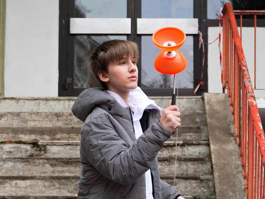 Ein Junge in grauer Jacke hält ein orangefarbenes Diabolo mit Stäben, auf einer Betontreppe mit orangem Geländer stehend.