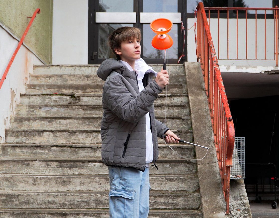 Ein Junge in grauer Jacke hält ein orangefarbenes Diabolo mit Stäben, auf einer Betontreppe mit orangem Geländer stehend.