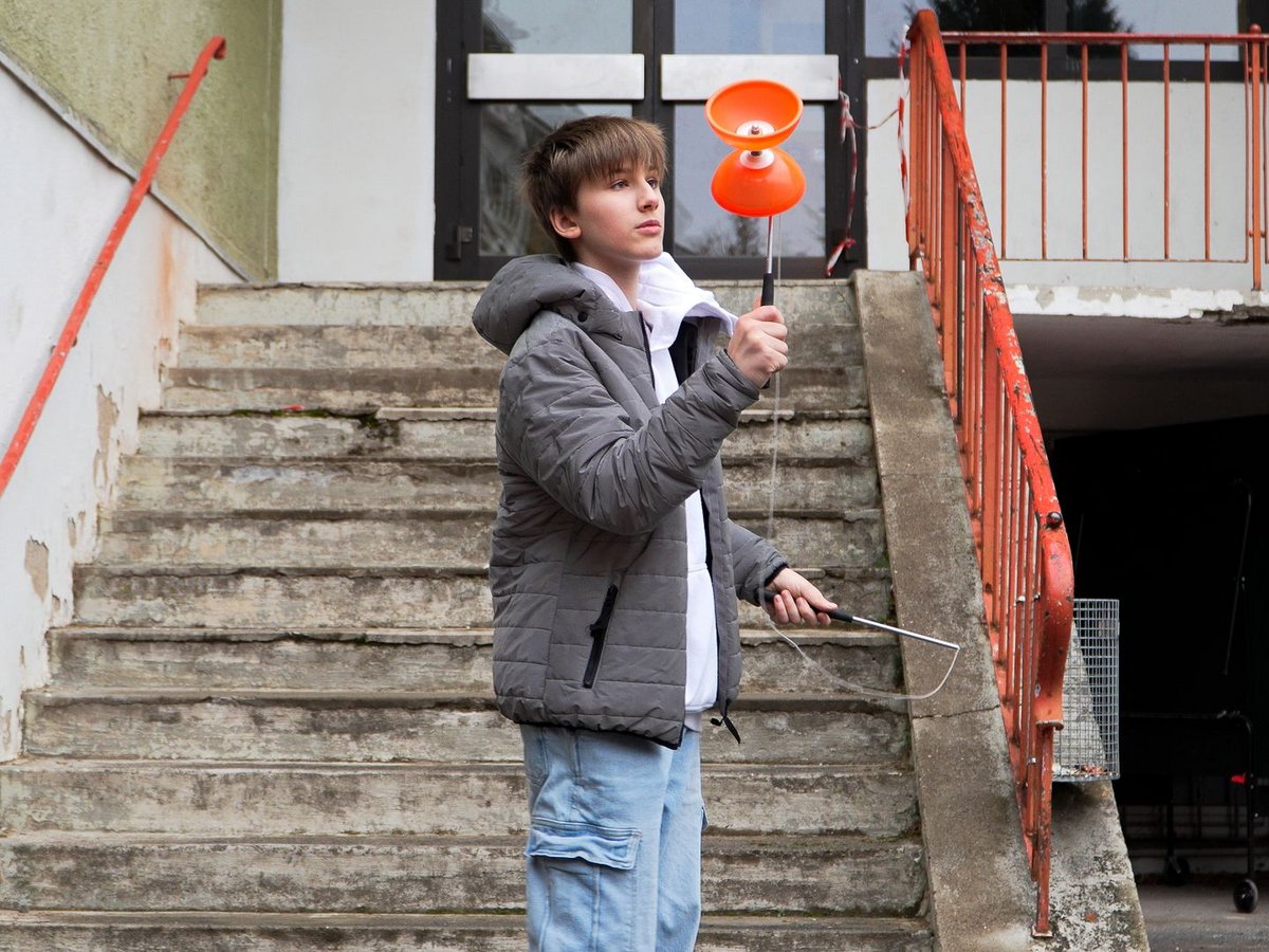 Ein Junge in grauer Jacke hält ein orangefarbenes Diabolo mit Stäben, auf einer Betontreppe mit orangem Geländer stehend.
