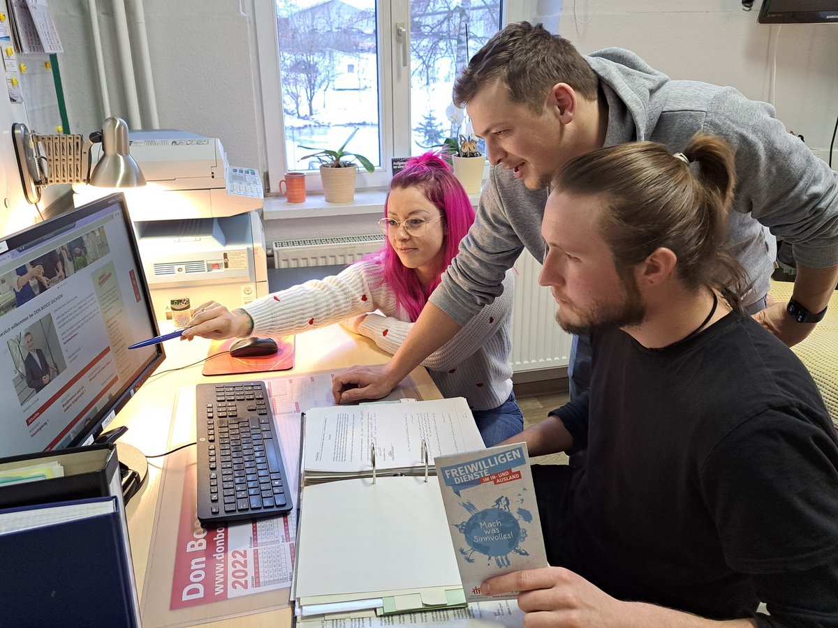 Drei junge Leute, eine mit pinken Haaren, betrachten einen Computerbildschirm und eine Broschüre über Freiwilligendienste im Büro.