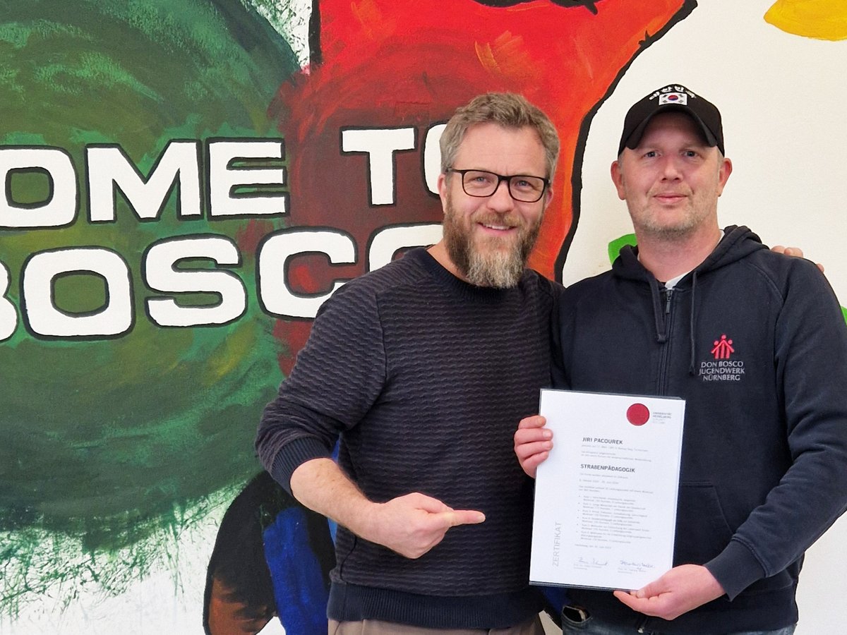 Zwei lächelnde Männer, einer hält ein Zertifikat für „Straßenpädagogik“, der andere zeigt darauf, vor einer bunten Wandmalerei.