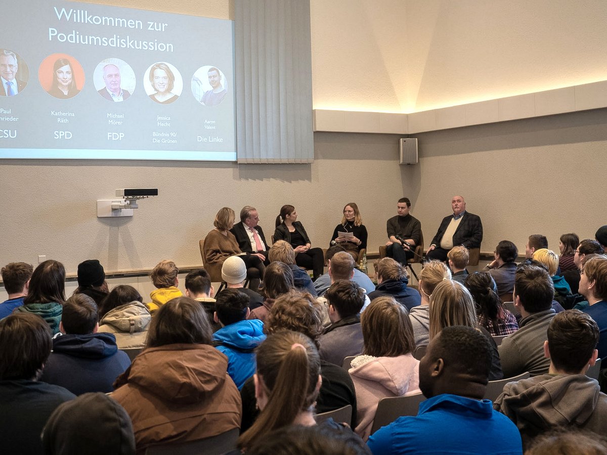 Eine politische Podiumsdiskussion mit fünf Rednern auf der Bühne, Namen/Parteien auf Leinwand, vor Publikum.