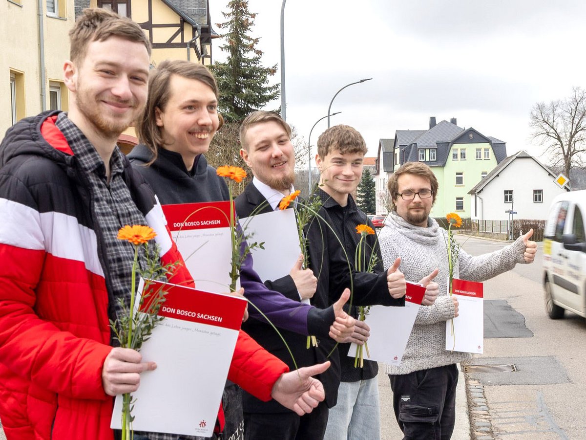 Fünf lächelnde junge Männer mit Don Bosco Zertifikaten und Blumen, die auf einer Straße Daumen hoch zeigen.