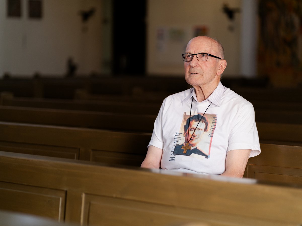 Älterer Mann mit Brille und T-Shirt mit religiösem Porträt und Kreuzkette, auf einer Kirchenbank sitzend.