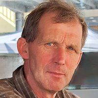 Nahaufnahme eines Mannes mittleren Alters mit blauen Augen und hellbraunem Haar, der ein braunes Karohemd trägt.