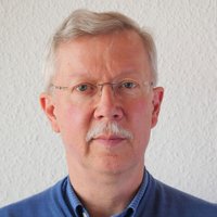 Nahaufnahme eines Mannes mit grauem Haar, Schnurrbart und randloser Brille, der ein blaues Hemd trägt.