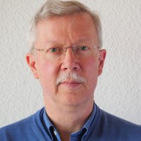 Nahaufnahme eines Mannes mit grauem Haar, Schnurrbart und randloser Brille, der ein blaues Hemd trägt.