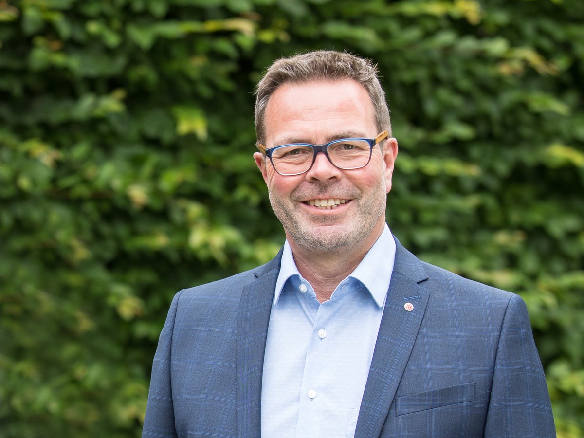 Lächelnder Mann mit grauen Haaren und blauer Brille, im karierten blauen Sakko, vor verschwommener grüner Hecke.