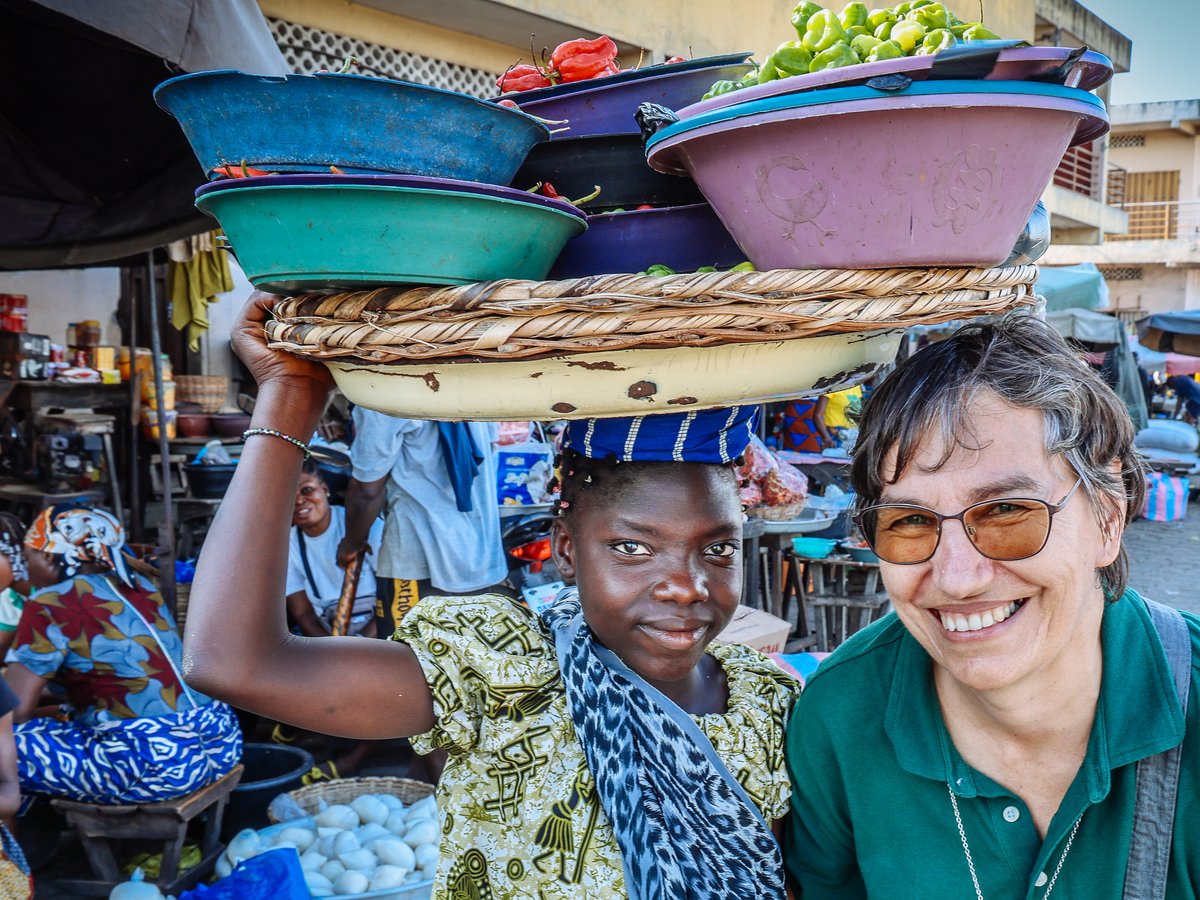 Zwei lächelnde Frauen, eine mit Gemüseschüsseln auf dem Kopf, auf einem belebten Markt.