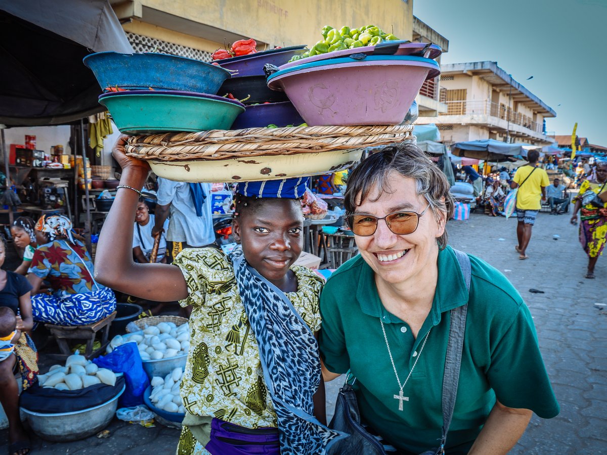 Zwei lächelnde Frauen, eine mit Gemüseschüsseln auf dem Kopf, auf einem belebten Markt.