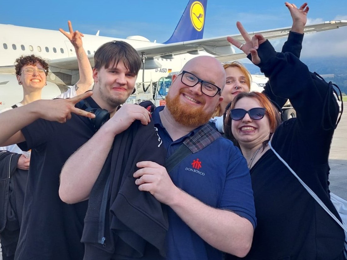 Fröhliche Gruppe junger Erwachsener und ein Mann im Don Bosco Hemd posieren mit Friedenszeichen vor einem Lufthansa Flugzeug.