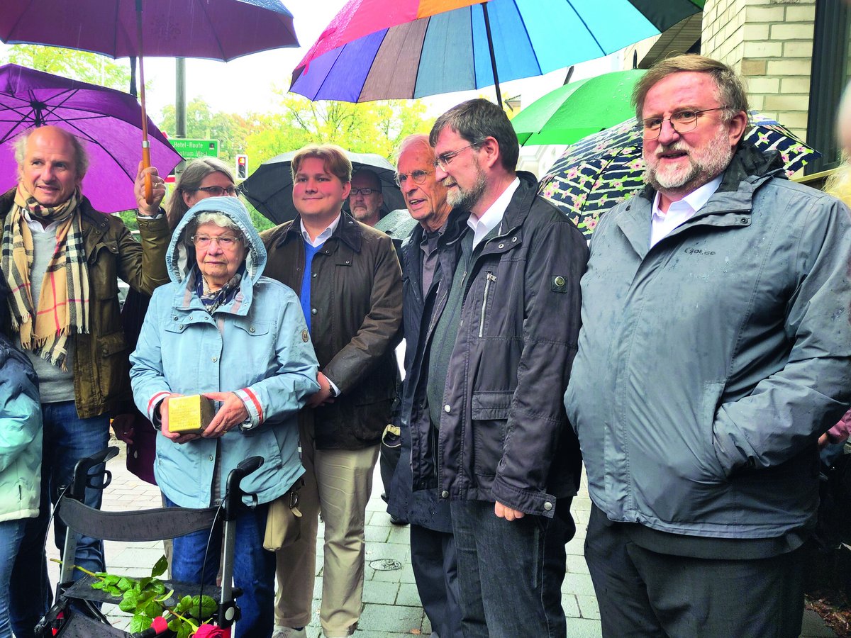 Gedenkveranstaltung im Regen: Eine ältere Frau hält einen Stolperstein, umgeben von einer Gruppe Menschen und Regenschirmen.