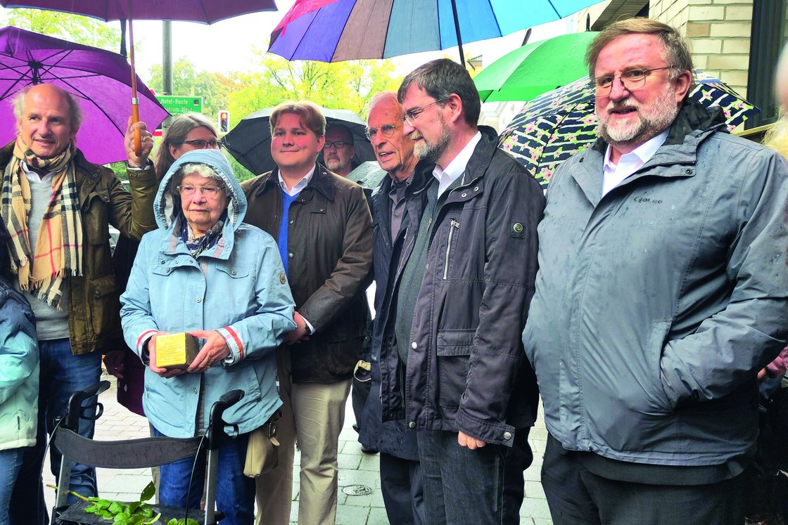 Gedenkveranstaltung im Regen: Eine ältere Frau hält einen Stolperstein, umgeben von einer Gruppe Menschen und Regenschirmen.