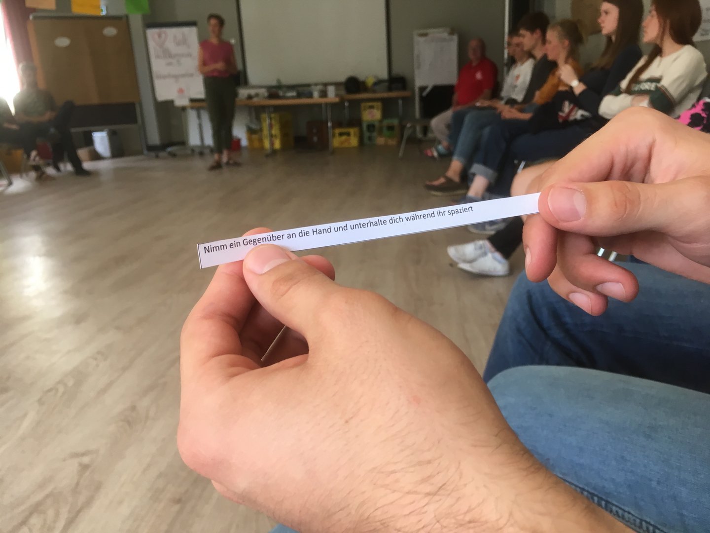 Hände halten Papierstreifen mit der Aufschrift: 'Nimm ein Gegenüber an die Hand und unterhalte dich während ihr spaziert'.