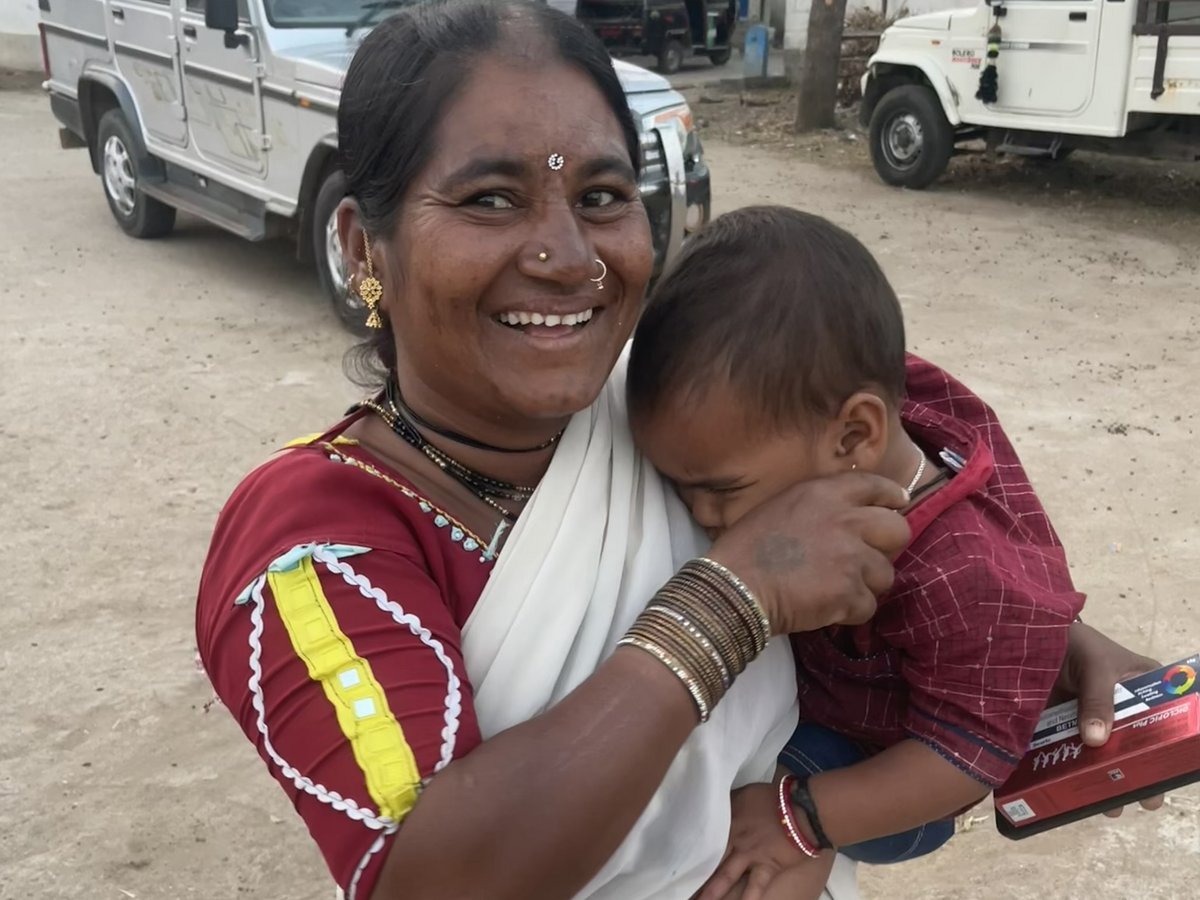 Eine strahlende indische Frau in traditioneller Kleidung hält ein Kind, mit Fahrzeugen und einer staubigen Straße im Hintergrund.