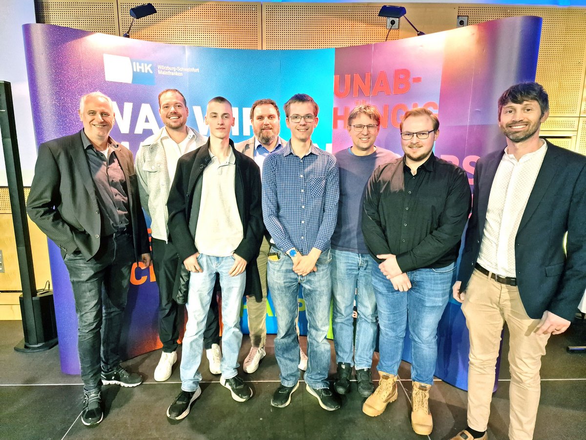 Acht lächelnde Männer posieren für ein Gruppenfoto vor einer Kulisse mit dem IHK Würzburg-Schweinfurt Mainfranken Logo.